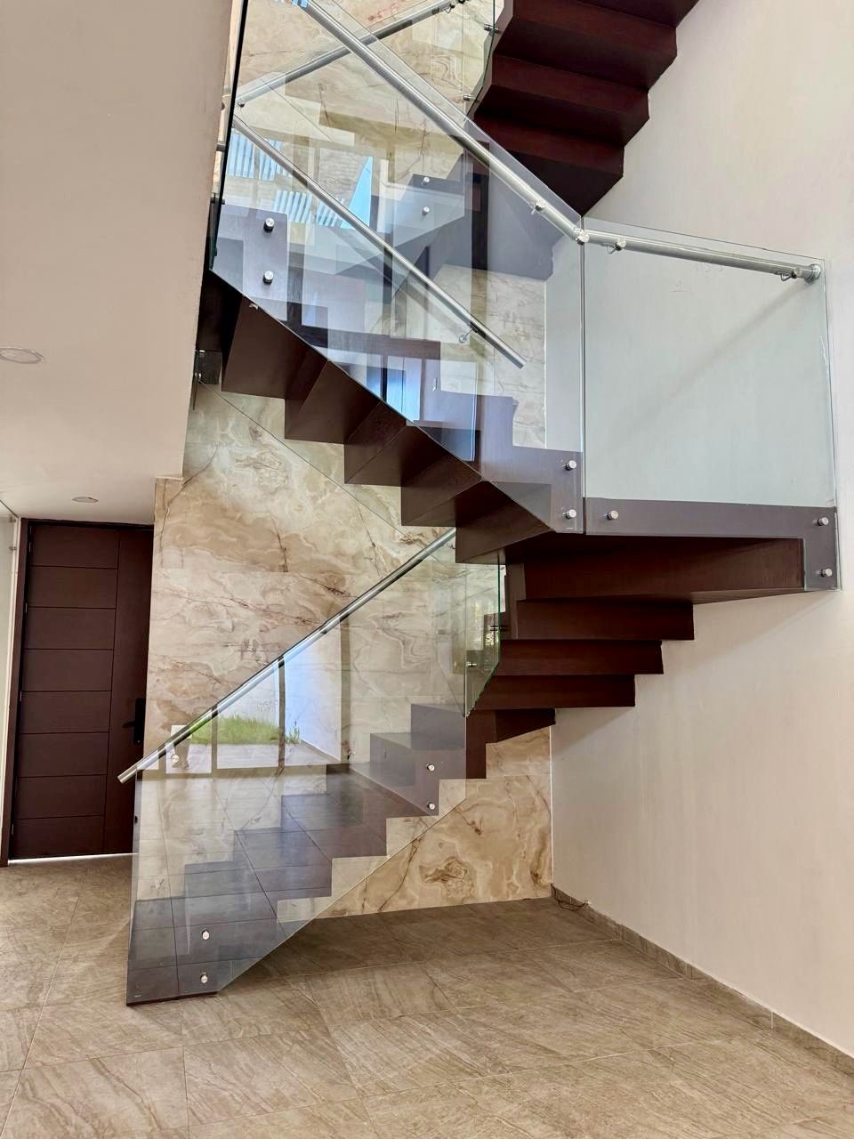 Escalera flotante de madera con barandal de cristal templado y muro de mármol a doble altura